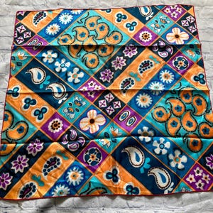 May include: A colorful square scarf with a geometric pattern. The design features paisley, floral, and abstract shapes in orange, teal, purple, and white, set against a dark blue background. The scarf has a red border.