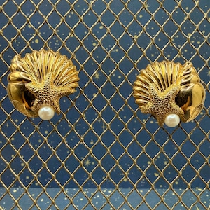 May include: A pair of gold-toned earrings shaped like scallop shells, each featuring a textured starfish overlay and a single white spherical pearl accent at the base. The earrings have a polished metallic finish and a detailed, ridged shell design.