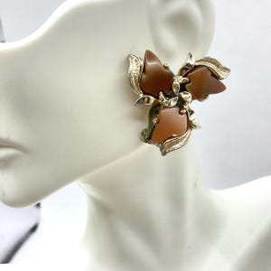 Puede incluir: Pendientes vintage de clip con diseño floral. Los pendientes tienen una base de metal dorado con tres pétalos marrones y detalles dorados. Los pendientes están sujetos a una cabeza de maniquí blanca.