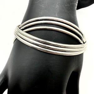 May include: A silver-toned bracelet with a crisscross design. The bracelet is composed of four rounded bands that intersect to create an eye-catching pattern. The jewelry is displayed on a black surface.