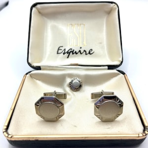 May include: A vintage Esquire cufflink and stud set in a black box with gold trim. The set includes two silver-toned cufflinks and a matching stud. The box is lined with cream-colored satin and features the Esquire logo.