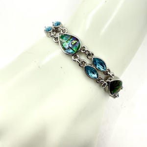 May include: Silver-toned bracelet featuring a series of decorative elements. The bracelet includes teardrop-shaped and oval-shaped iridescent abalone shells, and light blue, oval-shaped gemstones. The bracelet is on a white background.