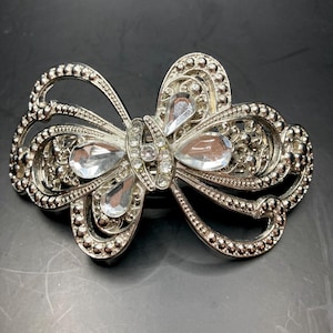 May include: A silver-toned hair clip shaped like a bow with clear rhinestones. The bow has a detailed design with small beads around the edges.