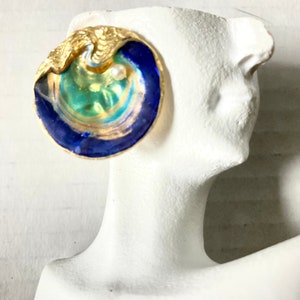 May include: A gold-toned seashell-shaped earring with a pearl accent. The earring features a swirling design of blue, teal, and gold enamel. The earring is displayed on a white mannequin head.