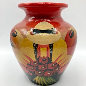 Puede incluir: Un jarrón de cerámica pintado a mano con un borde rojo y un diseño que presenta un sombrero, flores y formas abstractas en tonos de rojo, amarillo y negro. El jarrón tiene un acabado brillante.