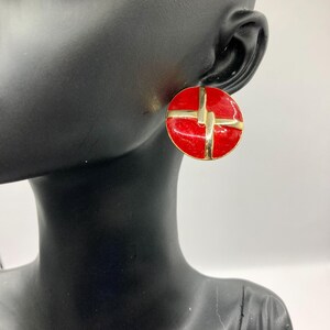 Op de afbeelding: Een paar rode en gouden clip-on oorbellen. De oorbellen zijn rond en hebben een geometrisch ontwerp.