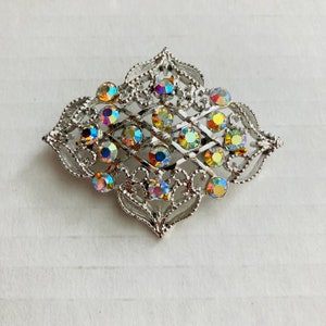 Puede incluir: Broche plateado con un diseño calado, con una serie de piedras preciosas redondas e iridiscentes. El broche tiene una forma geométrica simétrica con bordes decorativos. Las piedras preciosas reflejan un espectro de colores.