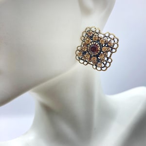 May include: A pair of gold-tone earrings with a square-shaped design. The earrings feature a cluster of small, clear rhinestones and a larger, dark purple rhinestone in the centre.