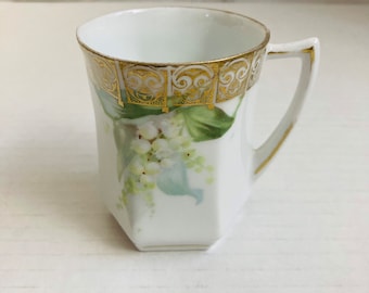 May include: White porcelain teacup with a gold rim and handle. The cup features a floral design of lily of the valley with green leaves and white flowers. The cup has a geometric shape.