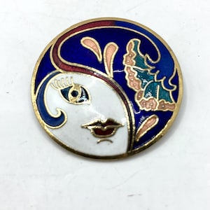 May include: Round, enameled brooch with a face design. The brooch features a white face with red lips, a blue eye, and a gold outline. The background is blue with pink and teal accents, and a gold border.