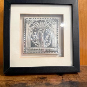 May include: A square, black-framed wall art piece featuring a white ceramic tile with a dark gray etched design. The tile depicts two arched panels containing Hebrew text and stylized palm fronds, flanked by two small Stars of David at the top.