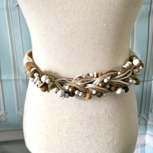 May include: A braided belt featuring a mix of white, tan, and metallic gold beads woven with natural jute twine and white cord. The belt displays a textured, multi-strand design with various bead shapes, including round, oval, and ribbed metallic accents.