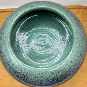 Puede incluir: Un cuenco redondo de cerámica con un exterior moteado de color verde azulado y un interior verde azulado más oscuro. El interior del cuenco presenta un diseño en espiral. El cuenco está sobre una superficie de madera.