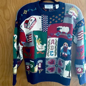 May include: A long-sleeve crew neck sweater featuring a multi-colored patchwork design with various knit motifs including a white goose, a sheep, the letters ABC, a house, and floral patterns in shades of navy blue, red, green, and white.