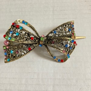 May include: A gold-toned metal hair clip in the shape of a bow. The bow features an openwork design and is embellished with colorful rhinestones in shades of red, blue, green, and pink. The clip has a barrette style closure.