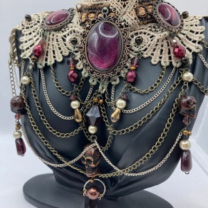 May include: An ornate, gothic-style necklace with a black base. The necklace features a large, oval, purple gemstone, surrounded by a bronze-colored filigree. It has multiple strands of chains, beads, and skull pendants, with a lace collar.