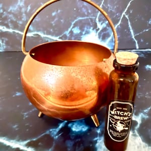 May include: A copper-colored metal cauldron with a curved handle and three small feet, accompanied by a brown glass bottle with a cork stopper. The bottle features a label that reads "SINCE 1692 WITCH'S BREW POTION NO 666" with a witch graphic.