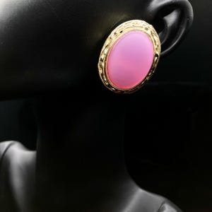 May include: A gold-toned oval earring with a light pink center. The earring has a textured border and is displayed on a black mannequin head. The earring is a vintage style.