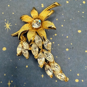 May include: A gold-tone metal brooch shaped like a flower with a textured finish. The center features a round clear rhinestone, while the petals and cascading floral elements are adorned with numerous marquise-cut clear rhinestones.