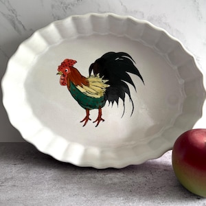 Może przedstawiać: Owalny ceramiczny talerz do serwowania w kolorze białym z falistym brzegiem. Talerz przedstawia kolorową ilustrację koguta z czerwonymi, zielonymi, żółtymi i czarnymi detalami. Czerwone jabłko znajduje się na pierwszym planie.