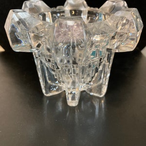 May include: A clear, faceted glass bowl with a starburst design. The bowl has a wide, open top and a geometric, multi-sided shape. The bowl is made of thick glass and has a reflective surface.