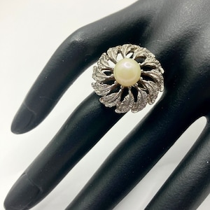 Könnte beinhalten: Ein silberner Ring mit einem großen weißen Perlmuttknopf in der Mitte. Der Ring ist mit kleinen Diamanten um die Perle besetzt und hat ein florales Design.