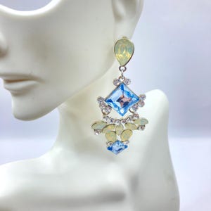 May include: A pair of dangle earrings with a light blue square-shaped gemstone surrounded by smaller clear rhinestones. The earrings also feature a teardrop-shaped light yellow opal gemstone at the top.