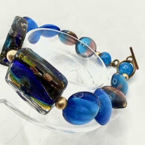 May include: A handcrafted bracelet featuring a mix of blue, gold, and iridescent glass beads. The bracelet includes rectangular and oval-shaped beads, connected by a bronze-colored clasp. The design incorporates various shades of blue and gold.
