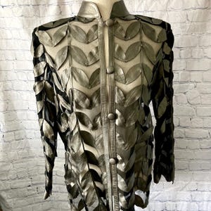 May include: A silver-toned jacket with a leaf design. The jacket features a high collar and is fastened with three buttons. The leaf pattern is layered over a sheer black fabric, creating a unique texture and visual depth. The jacket has long sleeves.