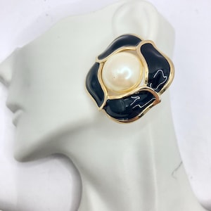 Op de afbeelding: Oorbellen in goudkleur met een zwart emaille bloemenontwerp en een witte parel in het midden.