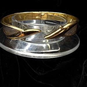 May include: A gold-toned hinged bracelet with a unique design. The bracelet features two stylized, textured bird-like elements at the opening. The bracelet is displayed on a clear, circular stand against a dark background.