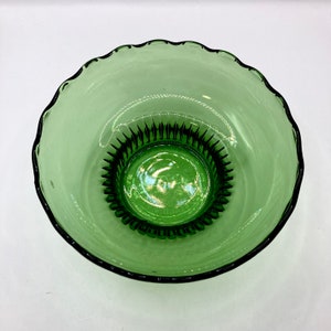 Puede incluir: Un cuenco de vidrio verde transparente con un borde festoneado. El cuenco tiene una base texturizada y un cuerpo liso y transparente. El cuenco está vacío y se encuentra sobre una superficie blanca.