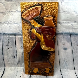 Può includere: Scultura a rilievo raffigurante una donna che trasporta una brocca e un cesto. L'opera presenta uno sfondo dorato strutturato. La donna indossa un abito rosso scuro con dettagli dorati. La scultura è un rettangolo verticale. Probabilmente è una decorazione murale.