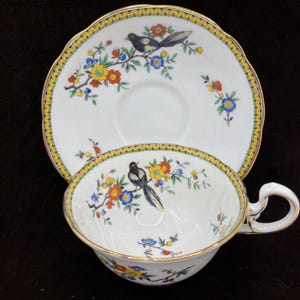 Puede incluir: Juego de taza y platillo blancos con un diseño floral y de pájaros de colores. La taza y el platillo tienen un borde festoneado con un borde amarillo y dorado. El diseño presenta un pájaro blanco y negro posado en una rama floreciente.