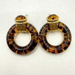 May include: A pair of vintage hoop earrings with a tortoiseshell pattern. The earrings have a gold-tone metal accent at the top and a post closure. The hoops are a translucent brown and yellow color.