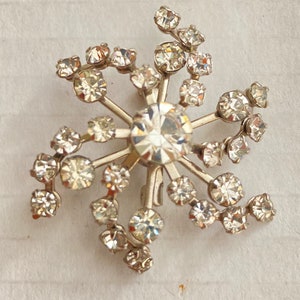 May include: A silver-toned brooch with a starburst design. The center features a large, clear, faceted stone, surrounded by radiating arms. Each arm has smaller, clear, round stones, creating a sparkling, elegant accessory.