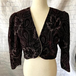Può includere: Bolero in velluto bordeaux scuro con finiture bianche. La giacca presenta un motivo di forme a vortice e disegni floreali con accenti argentati. Le maniche sono a tre quarti.