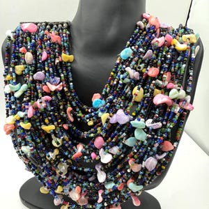 May include: A multi-strand necklace featuring numerous rows of small black glass beads interspersed with irregular, polished, pastel-colored shell or stone chips in shades of pink, yellow, blue, and white, creating a layered, textured appearance.