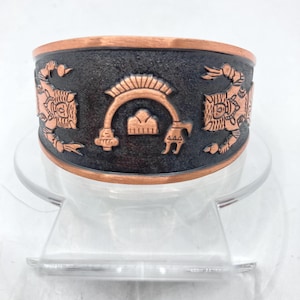 May include: A copper bracelet with a black oxidized finish. The bracelet features an embossed design of a stylized figure with a headdress and a crescent moon.