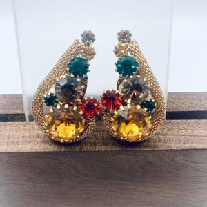 May include: A pair of gold tone clip-on earrings with a teardrop shape. The earrings are adorned with a cluster of colorful rhinestones in shades of green, red, and amber.