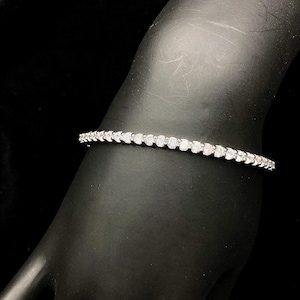 May include: A delicate silver bracelet with a row of sparkling, clear gemstones. The bracelet is set against a black background, highlighting its brilliance. The gemstones are evenly spaced, creating a continuous line of sparkle.