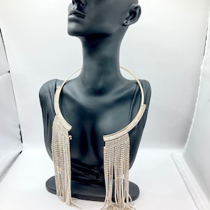 May include: A gold-toned collar necklace with a curved design, adorned with sparkling rhinestones. The necklace features a fringe of dangling rhinestones, creating a glamorous and eye-catching accessory. The necklace is displayed on a black mannequin.