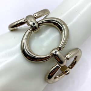 May include: A silver-toned metal bracelet with a large circular link and a T-bar closure. The bracelet is shown on a white surface, highlighting its polished finish and simple design. The bracelet is a piece of jewelry.