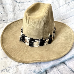 Könnte beinhalten: Beigefarbener Wildlederhut mit breiter Krempe und hohem Kopf. Der Hut hat ein dekoratives Band aus schwarz-weißen Federn am unteren Rand des Kopfes. Der Hut ist neutralfarben und scheint ungetragen zu sein.