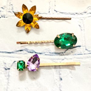 May include: Three gold hair clips with different coloured rhinestones. One clip has a yellow flower-shaped rhinestone, another has a green oval rhinestone, and the last has a green square and a purple teardrop rhinestone.