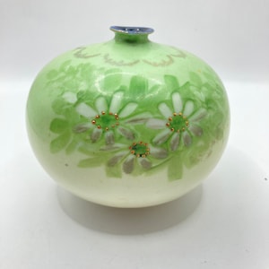 May include: A small, round, green ceramic vase with white flowers and gold accents. The vase has a narrow neck and a wide base.
