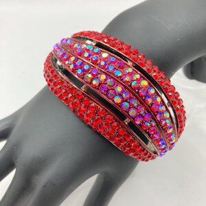 Puede incluir: Una pulsera brazalete roja con filas de pequeños pedrería rojos e iridiscentes brillantes. La pulsera es de metal y tiene un diseño ranurado. Las piedras preciosas reflejan múltiples colores.