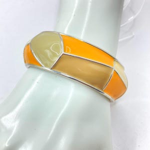 Könnte beinhalten: Ein silberfarbenes Metallarmband mit geometrischem Design. Das Armband hat orange, beige und cremefarbene Emaille-Abschnitte. Das Armband hat einen Durchmesser von etwa 6,35 cm.