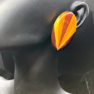 May include: An oval wooden earring with a geometric design. The earring features dark brown, orange, and yellow wood pieces arranged in a fan-like pattern. The earring is attached to a black mannequin head.