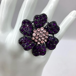 Puede incluir: Un anillo grande con forma de flor morada con un centro de piedras de imitación rosas. El anillo está hecho de metal y tiene un acabado negro.